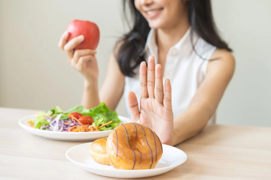happy asian young woman's hand denies sweet donut on plate, and looks at red apple and green vegetable salad,